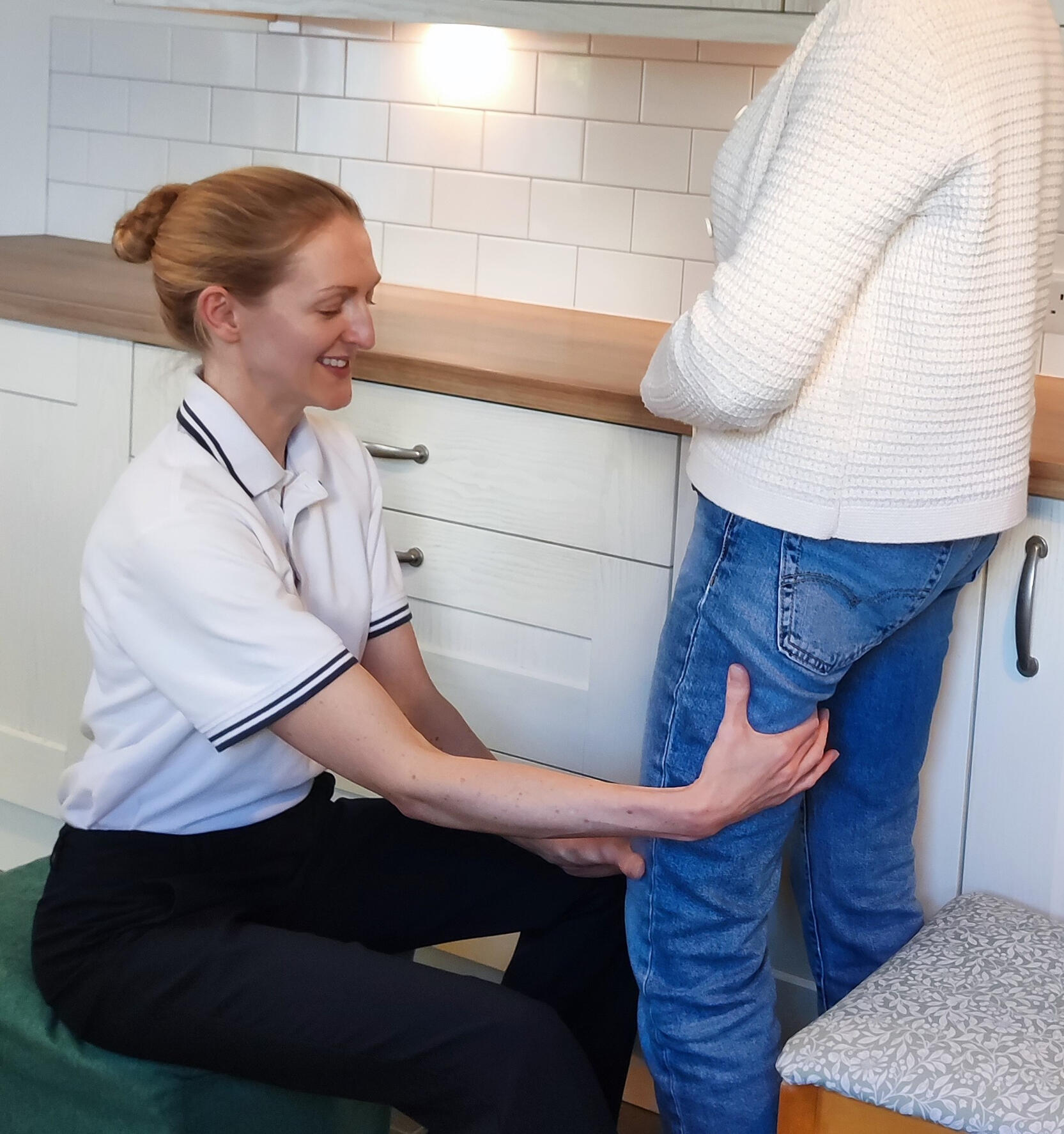 Physio on Leg Physiotherapist assisting a person standing in a kitchen, supporting their thigh while guiding a leg movement during a rehabilitation session.